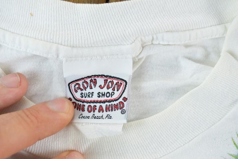 Puede incluir: Primer plano de una etiqueta de camiseta blanca de Ron Jon Surf Shop. La etiqueta es rectangular con un logotipo rojo y negro que dice "Ron Jon Surf Shop" y "One of a Kind". La etiqueta tambi&eacute;n incluye la ubicaci&oacute;n "Cocoa Beach, Fla."
