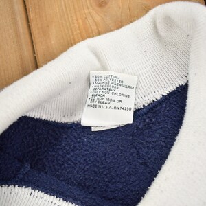 May include: A white tag with blue text detailing the care instructions for a navy blue and white striped sweatshirt. The tag reads: 50% Cotton/ 50% Polyester, Machine Wash Warm, Wash Colors Separately, Only Non-Chlorine Bleach, Do Not Iron or Dry Clean, Made in USA. RN 74230.