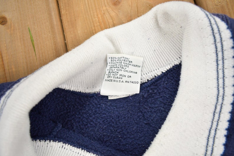 May include: A white tag with blue text detailing the care instructions for a navy blue and white striped sweatshirt. The tag reads: 50% Cotton/ 50% Polyester, Machine Wash Warm, Wash Colors Separately, Only Non-Chlorine Bleach, Do Not Iron or Dry Clean, Made in USA. RN 74230.