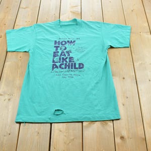 Peut inclure: Un t-shirt à manches courtes de couleur bleu sarcelle vif avec un texte bleu foncé sur le devant qui indique How To Eat Like A Child, accompagné d'un texte plus petit et d'un petit trou près de l'ourlet inférieur du tissu du vêtement.