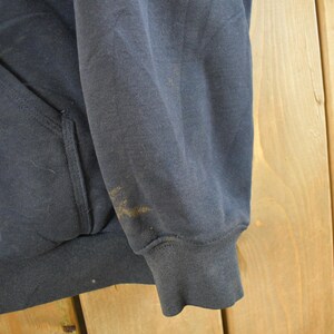 Puede incluir: Primer plano de una sudadera con capucha azul marino. La imagen muestra la manga y la zona del bolsillo de la prenda. Hay algunas manchas en la manga. La sudadera parece estar hecha de un material suave y desgastado.