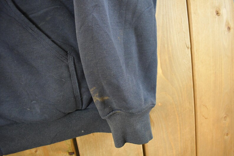 Puede incluir: Primer plano de una sudadera con capucha azul marino. La imagen muestra la manga y la zona del bolsillo de la prenda. Hay algunas manchas en la manga. La sudadera parece estar hecha de un material suave y desgastado.