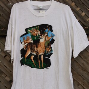 Pode incluir: Camiseta branca com uma estampa gr&aacute;fica de um veado de cauda branca em um ambiente de floresta. O veado est&aacute; cercado por folhagem verde e c&eacute;u azul. O texto "Jim Shore" est&aacute; impresso no canto inferior direito do gr&aacute;fico.