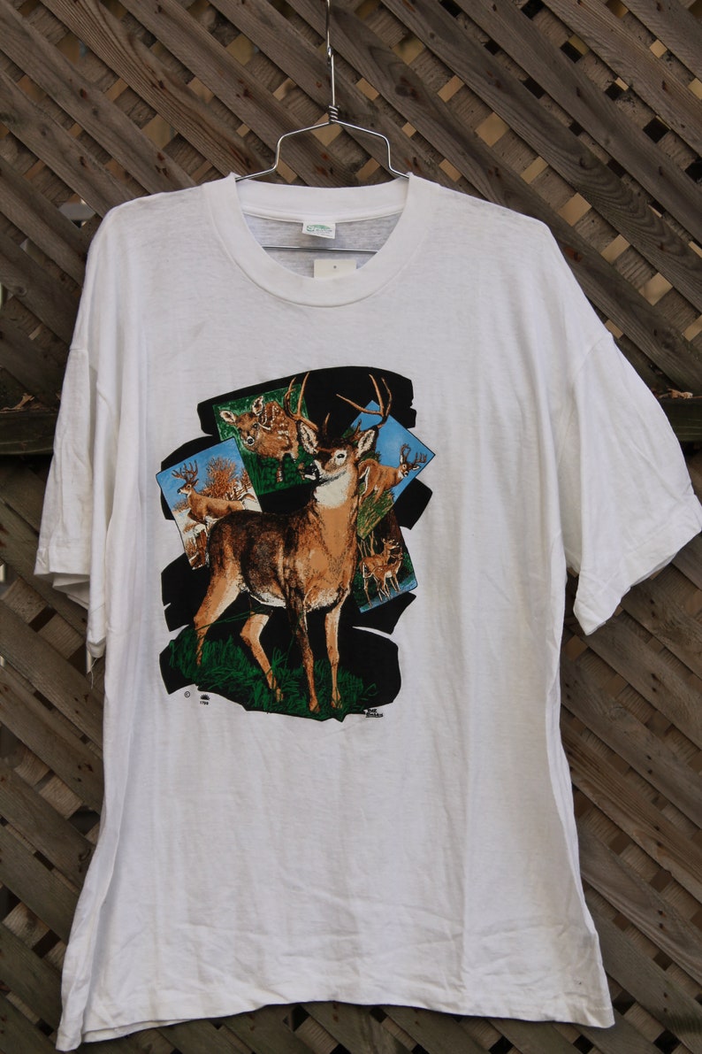 Pode incluir: Camiseta branca com uma estampa gr&aacute;fica de um veado de cauda branca em um ambiente de floresta. O veado est&aacute; cercado por folhagem verde e c&eacute;u azul. O texto "Jim Shore" est&aacute; impresso no canto inferior direito do gr&aacute;fico.