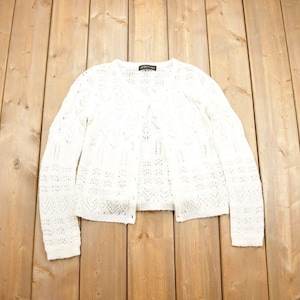 May include: A white, open-front cardigan with a delicate, open-knit design. The long-sleeved sweater has a button closure and a textured pattern. The garment is displayed on a light brown wooden surface.