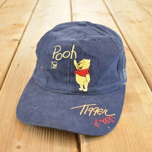 May include: Navy blue baseball cap featuring embroidered Winnie the Pooh characters. The cap has the word "Pooh" above an image of Pooh Bear, and the word "Tigger" below. The design includes a red and yellow color scheme.