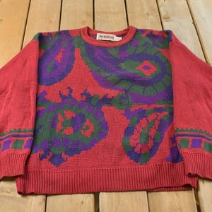 Puede incluir: Un suéter de manga larga con cuello redondo en un tono rojo intenso que presenta un llamativo patrón de cachemira en morado y verde en el pecho. La prenda incluye puños y dobladillo acanalados, con un borde geométrico decorativo sobre los puños.