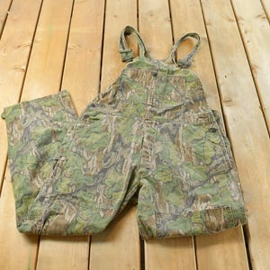 May include: Camouflage-patterned overalls with adjustable straps. The green, brown, and tan print covers the entire garment, including the bib, pockets, and legs. The overalls are made of a durable fabric, suitable for outdoor activities.