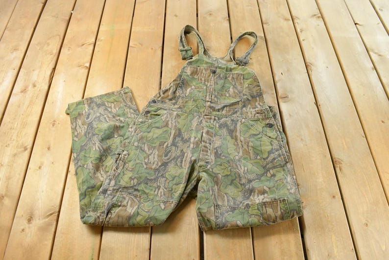 May include: Camouflage-patterned overalls with adjustable straps. The green, brown, and tan print covers the entire garment, including the bib, pockets, and legs. The overalls are made of a durable fabric, suitable for outdoor activities.