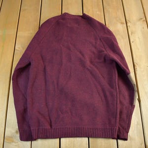 Puede incluir: Un su&eacute;ter burdeos con cuello redondo y mangas largas. El su&eacute;ter tiene un dobladillo y pu&ntilde;os acanalados. La parte trasera del su&eacute;ter es visible y el material parece ser un tejido suave. El su&eacute;ter es de color liso con un dise&ntilde;o sencillo.