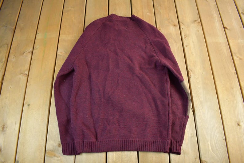 Puede incluir: Un su&eacute;ter burdeos con cuello redondo y mangas largas. El su&eacute;ter tiene un dobladillo y pu&ntilde;os acanalados. La parte trasera del su&eacute;ter es visible y el material parece ser un tejido suave. El su&eacute;ter es de color liso con un dise&ntilde;o sencillo.