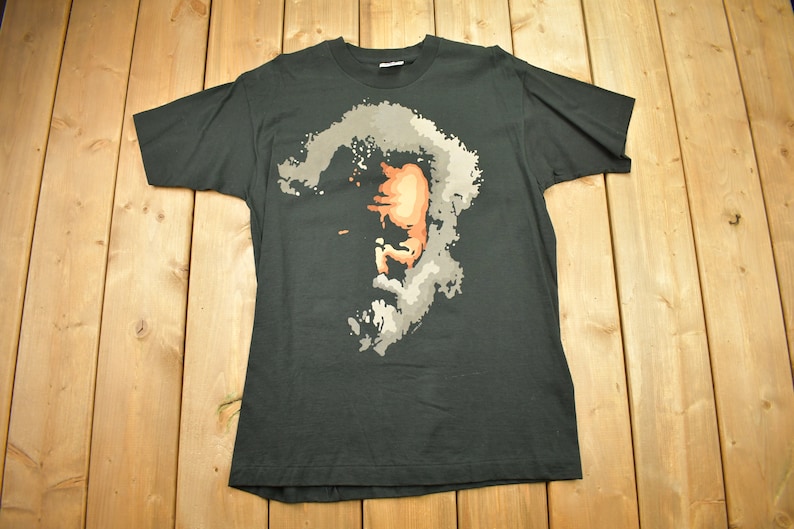 May include: A black t-shirt with a faded, pixelated image of a man's face in shades of gray, orange, and white.