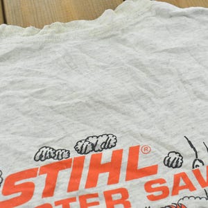 Op de afbeelding: Een lichtgrijs t-shirt met korte mouwen met de oranje tekst STIHL en de gedeeltelijke zwarte tekst TER SAW. Het ontwerp bevat zwart-witte grafische illustraties van wolken of rookpluimen rondom de dikgedrukte oranje belettering.