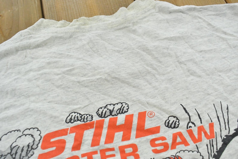 Op de afbeelding: Een lichtgrijs t-shirt met korte mouwen met de oranje tekst STIHL en de gedeeltelijke zwarte tekst TER SAW. Het ontwerp bevat zwart-witte grafische illustraties van wolken of rookpluimen rondom de dikgedrukte oranje belettering.