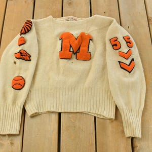 May include: Cream-coloured vintage sweater with orange and black accents. Features an orange "M" on the chest, sports-themed patches on the sleeves, and the number "55". Ribbed cuffs and hem complete the look.
