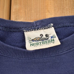Puede incluir: Una sudadera azul marino con una etiqueta blanca que dice "Northern Reflections" y presenta dos p&aacute;jaros buceadores.