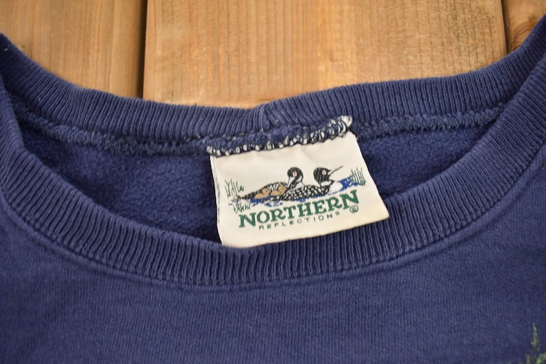 Puede incluir: Una sudadera azul marino con una etiqueta blanca que dice "Northern Reflections" y presenta dos p&aacute;jaros buceadores.