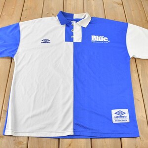 May include: A white and blue polo shirt with a split design. The left side is white with a small Umbro logo. The right side is blue with a large "Umbro" logo and the text "Blue - The future and the truth."