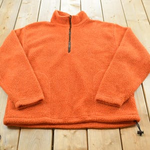 May include: An orange fleece pullover with a black zip and a high collar. The long-sleeved top has a drawstring at the bottom and two front pockets. The soft, textured material is spread out on a wooden surface.