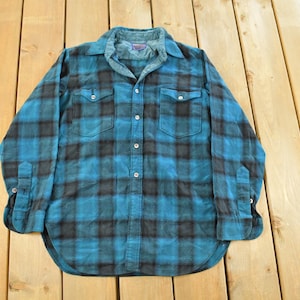 May include: A blue and black plaid flannel shirt with long sleeves, two chest pockets, and button closures. The shirt is laid flat on a wooden surface. The collar and cuffs are a lighter shade of blue.