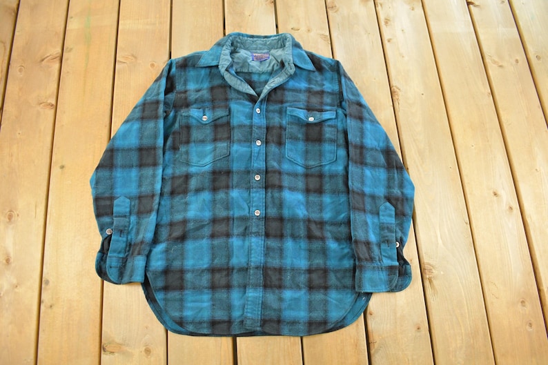 May include: A blue and black plaid flannel shirt with long sleeves, two chest pockets, and button closures. The shirt is laid flat on a wooden surface. The collar and cuffs are a lighter shade of blue.