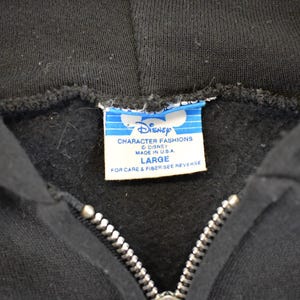 May include: Close-up of a black hooded sweatshirt with a silver zipper. The Disney Character Fashions label is visible, indicating the brand and size "LARGE". Made in the USA.