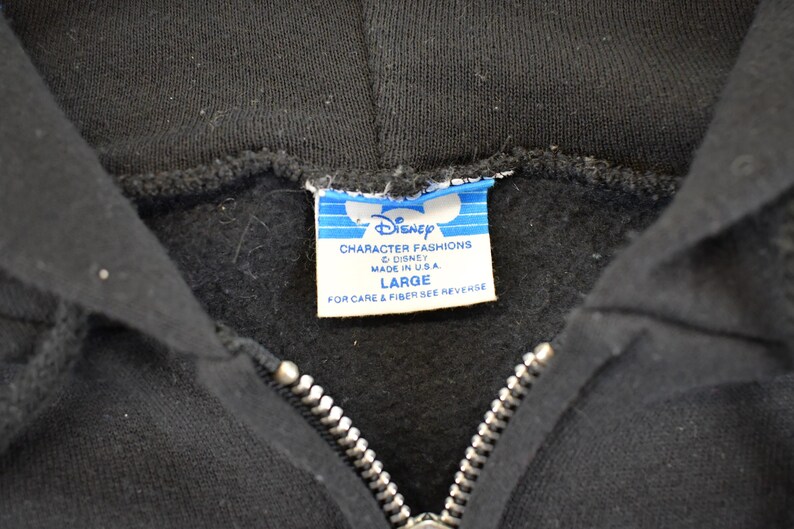 May include: Close-up of a black hooded sweatshirt with a silver zipper. The Disney Character Fashions label is visible, indicating the brand and size "LARGE". Made in the USA.