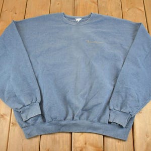 May include: A light blue Champion sweatshirt with a crew neck and long sleeves. The sweatshirt has a small Champion logo on the chest. The garment is made of a soft material and is laid out on a wooden surface.