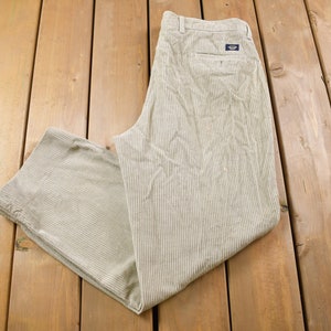 May include: Beige corduroy trousers with thin white stripes. The trousers have a single back pocket with a small logo patch.