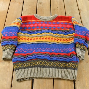 Könnte beinhalten: Ein farbenfroher Pullover mit Muster und Rundhalsausschnitt. Der Pullover hat horizontale Bänder in Rot, Blau, Gelb und Grau mit verschiedenen geometrischen und floralen Designs. Die Bündchen und der Saum sind grau.