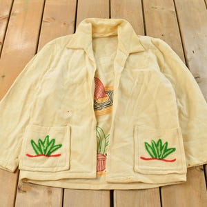 Peut inclure: Veste crème avec des broderies de cactus et de motifs floraux. La veste comporte deux poches avant avec des cactus brodés verts et rouges, et des motifs floraux supplémentaires sur les manches. Le devant ouvert révèle une impression colorée.
