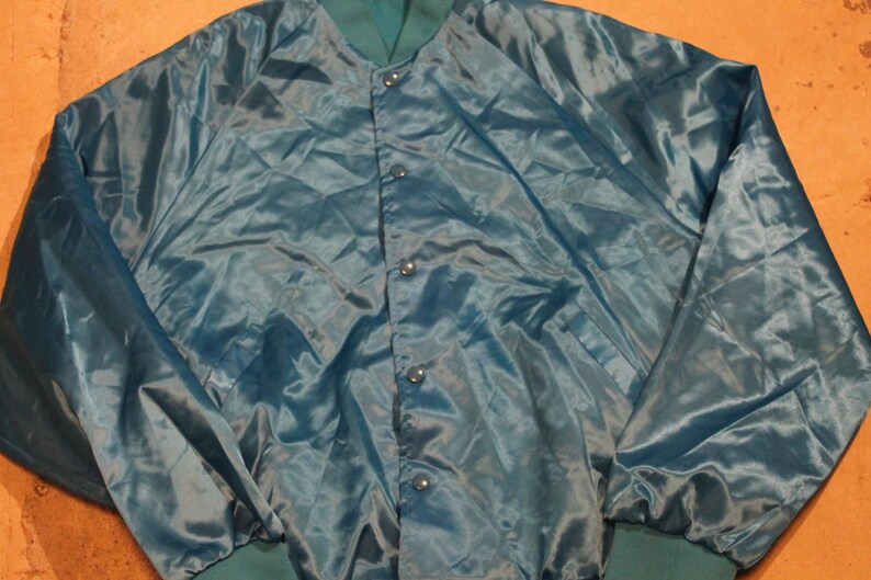 May include: Teal satin jacket with a button-up front and green ribbed cuffs and collar. The jacket has a shiny, smooth texture and a relaxed fit. The jacket is a vintage style.