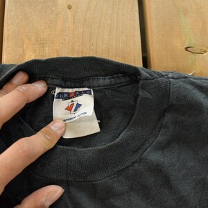 Op de afbeelding: Close-up van een zwarte t-shirtkraag met een label. Het label is wit met rode en blauwe accenten en de merknaam "JERZEES". Het shirt ligt op een houten oppervlak.