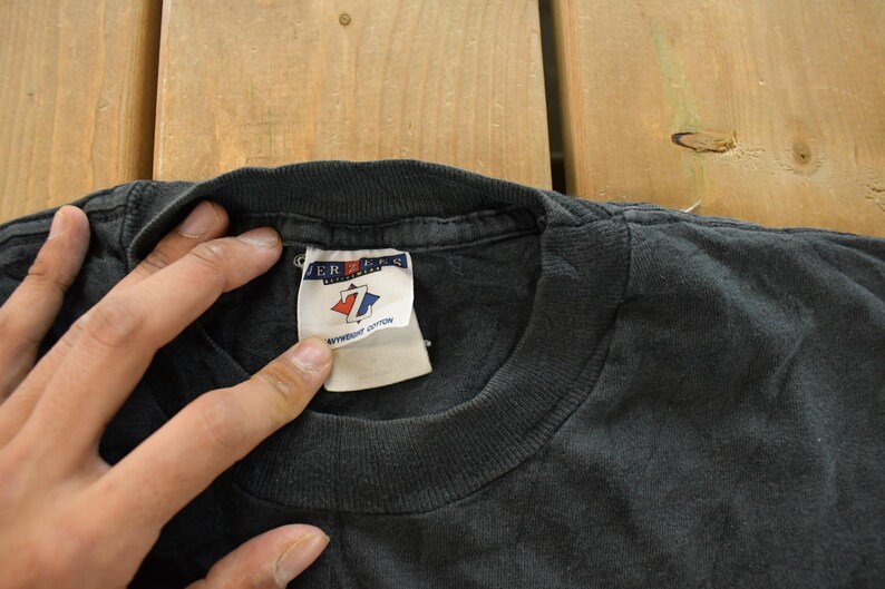 Op de afbeelding: Close-up van een zwarte t-shirtkraag met een label. Het label is wit met rode en blauwe accenten en de merknaam "JERZEES". Het shirt ligt op een houten oppervlak.