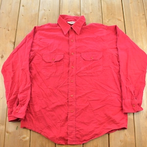 May include: A long-sleeved button-down shirt in a solid bright red colour. The shirt features a standard collar, a full-length front button placket, and two chest pockets with buttoned flaps. The cuffs are also finished with matching red buttons.
