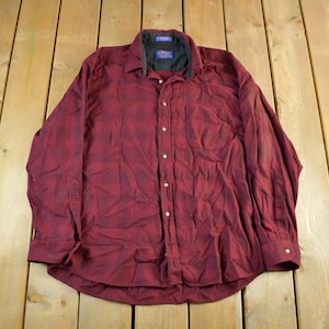 Puede incluir: Una camisa abotonada de manga larga en color burdeos oscuro con un sutil estampado de cuadros. La camisa presenta un cuello clásico, un bolsillo en el pecho y puños abotonados. El cuello tiene un forro negro.