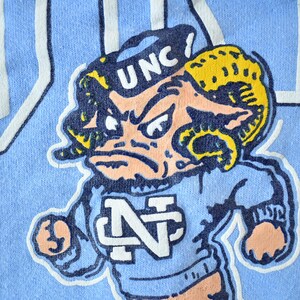 May include: A blue t-shirt with a cartoon mascot wearing a blue and white "UNC" hat and a blue and white "N" shirt. The mascot has a yellow and blue ram horn and a pink face.