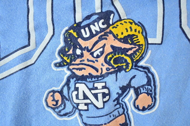 May include: A blue t-shirt with a cartoon mascot wearing a blue and white "UNC" hat and a blue and white "N" shirt. The mascot has a yellow and blue ram horn and a pink face.