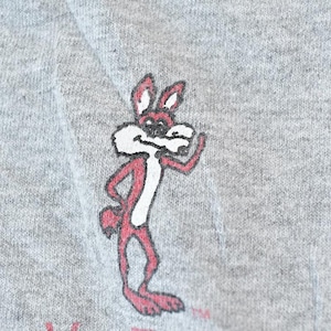 Peut inclure: Un sweat-shirt gris avec un personnage de lapin de dessin animé en rouge et blanc. Le lapin est debout, une main sur la hanche et l'autre levée en signe de salut. Le motif est légèrement délavé.