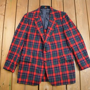 May include: A tartan blazer in red, green, and navy blue. The jacket features a classic two-button fastening, notched lapels, and two front pockets. The lining is a solid navy blue. The blazer is displayed on a wooden surface.
