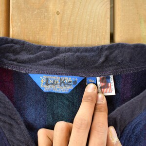 Puede incluir: Primer plano de una prenda de lana Pendleton con cuello azul marino y un estampado de rayas en tonos morado, azul y verde. La prenda tiene una etiqueta azul con texto blanco y una peque&ntilde;a etiqueta con la bandera americana.