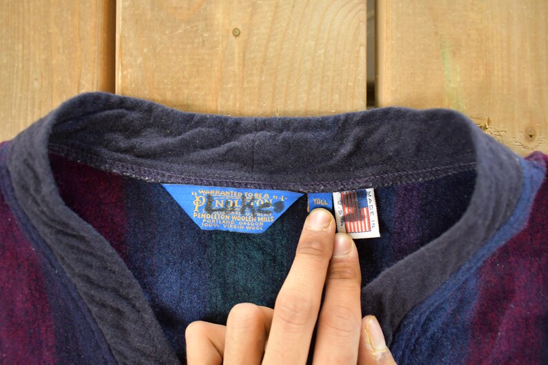 Puede incluir: Primer plano de una prenda de lana Pendleton con cuello azul marino y un estampado de rayas en tonos morado, azul y verde. La prenda tiene una etiqueta azul con texto blanco y una peque&ntilde;a etiqueta con la bandera americana.