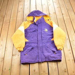May include: A purple and yellow hooded winter jacket with a team logo on the front. The logo is a Viking helmet with horns and the text "TM".