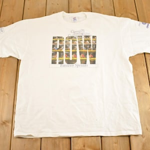 May include: White t-shirt with a graphic featuring the word "ROW" in large letters, with a picture of a rowing competition inside the letters. The text "Champions of Champions Eastern Sprints" is above and below the word "ROW".