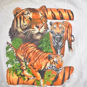 K&ouml;nnte beinhalten: Wei&szlig;es T-Shirt mit einem grafischen Druck von drei Tigern. Die Tiger befinden sich in einer Dschungelumgebung mit gr&uuml;nen Bl&auml;ttern und Ranken. Die Tiger sind orange und schwarz mit wei&szlig;en Streifen.
