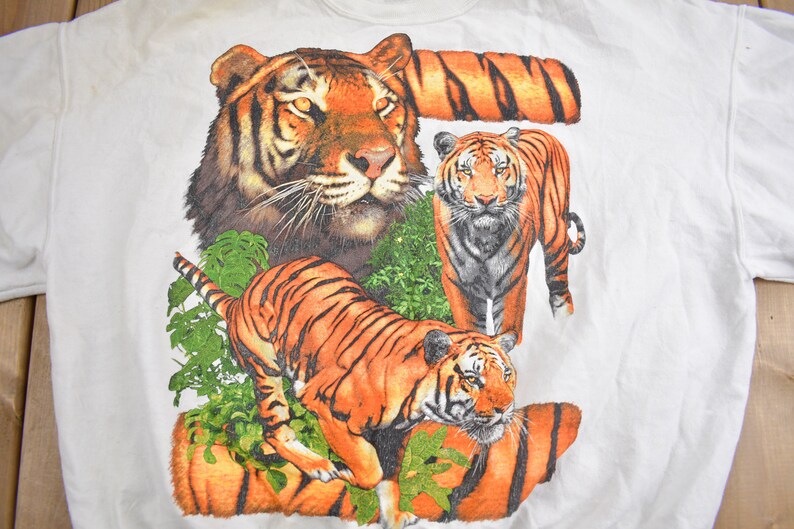 K&ouml;nnte beinhalten: Wei&szlig;es T-Shirt mit einem grafischen Druck von drei Tigern. Die Tiger befinden sich in einer Dschungelumgebung mit gr&uuml;nen Bl&auml;ttern und Ranken. Die Tiger sind orange und schwarz mit wei&szlig;en Streifen.