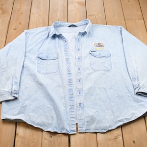 Puede incluir: Una camisa vaquera azul claro de manga larga y cierre de botones. La camisa tiene dos bolsillos en el pecho y presenta un logotipo bordado que dice "Cat's Meow" con un gráfico de gato.