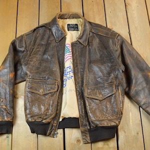 May include: A vintage brown leather bomber jacket with two front pockets and a black ribbed hem and cuffs. The jacket has a distressed appearance and a light-colored lining with text and an American flag graphic.