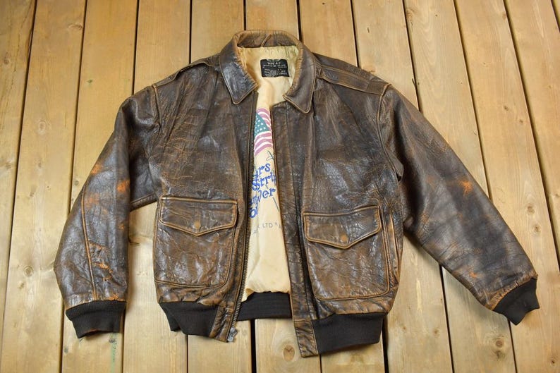 May include: A vintage brown leather bomber jacket with two front pockets and a black ribbed hem and cuffs. The jacket has a distressed appearance and a light-colored lining with text and an American flag graphic.