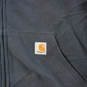 Puede incluir: Primer plano de una sudadera con capucha Carhartt azul marino con cremallera. La sudadera con capucha tiene un bolsillo delantero y una peque&ntilde;a etiqueta cuadrada con el logotipo de Carhartt en naranja y blanco. La cremallera es visible en el centro.